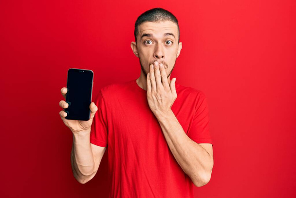Hispanic young man holding smartphone showing blank screen covering mouth with hand, shocked and afraid for mistake. surprised expression  - Valokuva, kuva