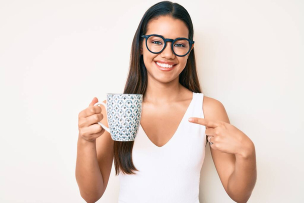 jong mooi latijn meisje dragen bril houden koffie lachend gelukkig wijzend met hand en vinger  - Foto, afbeelding