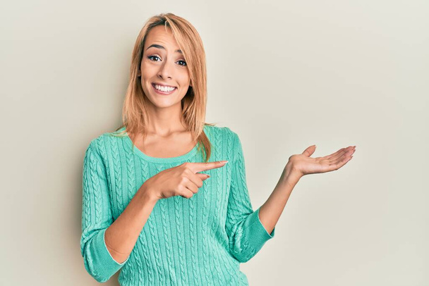 Beautiful blonde woman wearing casual winter sweater amazed and smiling to the camera while presenting with hand and pointing with finger.  - Foto, Imagem