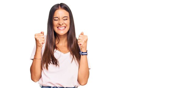 Gündelik beyaz tişört giyen genç İspanyol kadın başarı için heyecanlanıyor kolları havada ve gözleri kapalı zaferi kutluyor. Kazanan konsept.  - Fotoğraf, Görsel