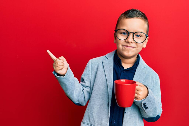 Malý chlapec nosí brýle, pije z červeného hrnku, usmívá se a šťastně ukazuje rukou a prstem na stranu.  - Fotografie, Obrázek