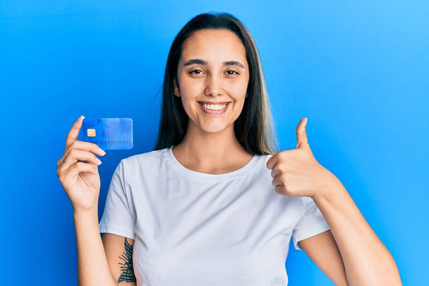 Giovane donna ispanica in possesso di carta di credito sorridente felice e positivo, pollice in su facendo eccellente e segno di approvazione  - Foto, immagini