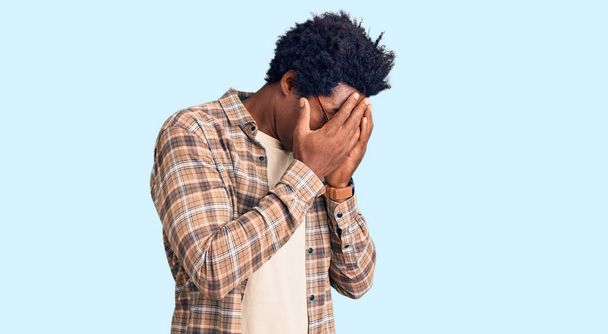 Schöner afrikanisch-amerikanischer Mann mit Afrohaaren, lässiger Kleidung und Brille mit traurigem Gesichtsausdruck, der beim Weinen das Gesicht mit den Händen bedeckt. Depressionskonzept.  - Foto, Bild