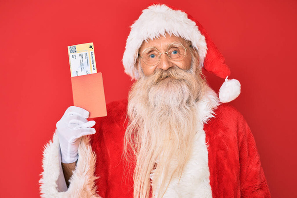 Viejo hombre mayor con el pelo gris y la barba larga con traje de Santa Claus sosteniendo tarjeta de embarque mirando positiva y feliz de pie y sonriendo con una sonrisa confiada mostrando los dientes  - Foto, Imagen