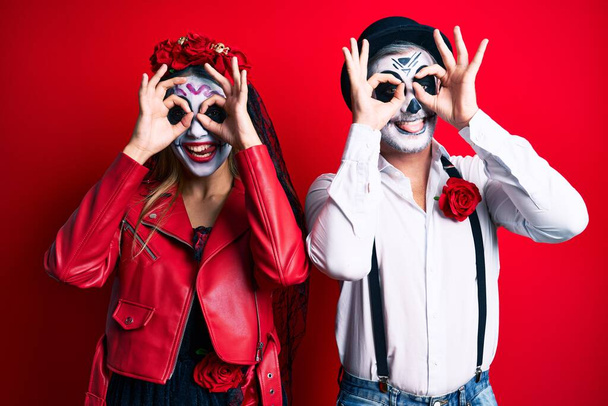 Couple wearing day of the dead costume over red doing ok gesture like binoculars sticking tongue out, eyes looking through fingers. crazy expression.  - Photo, Image