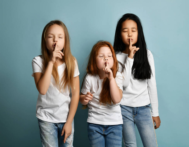 Multiracial girls friends in jeans and comfortable longsleeves standing showing silence sign with fingers and eyes closed - Foto, Bild