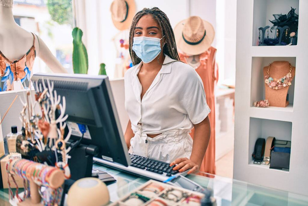 Joven mujer afroamericana sonriendo feliz trabajando en la caja con la máscara de seguridad coronavirus en la tienda al por menor - Foto, imagen