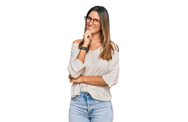 Jovem mulher vestindo roupas casuais e óculos olhando confiante para a câmera com sorriso com braços cruzados e mão levantada no queixo. pensamento positivo.  - Foto, Imagem