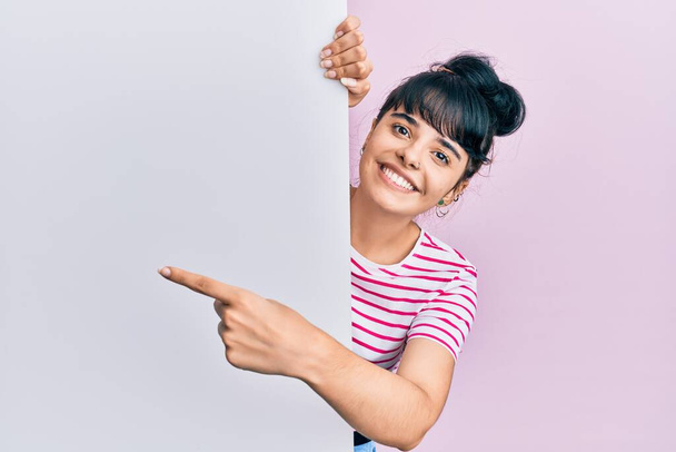 Młoda latynoska dziewczyna trzyma pusty sztandar uśmiechając się szczęśliwy wskazując ręką i palcem  - Zdjęcie, obraz