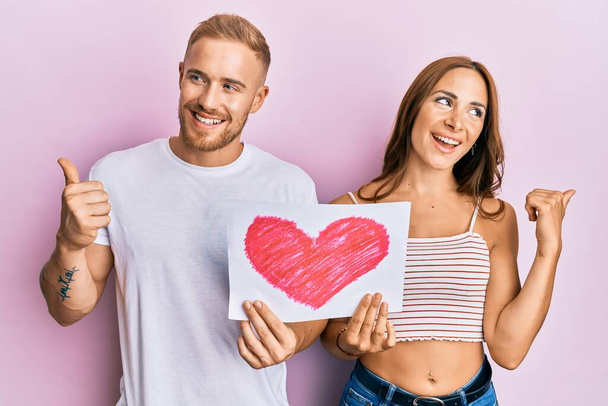 Молодая пара подружек и бойфрендов, держащих в руках сердечный рисунок, указывая пальцем вверх в сторону, улыбаясь счастливо с открытым ртом  - Фото, изображение