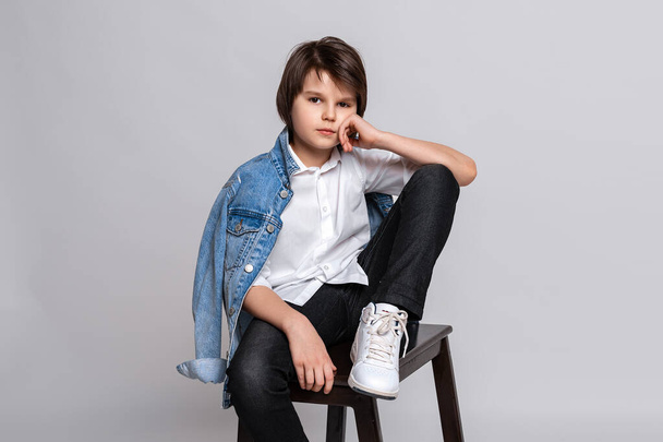 Handsome fashion little boy 11-13 years old sits on a chair in studio. Dressed in a white shirt, black jeans, jean jacket and white sneakers. Model Test. Fashion and people concept - Foto, imagen