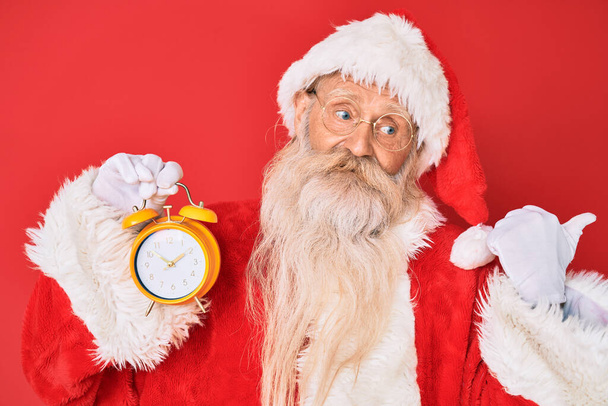 Старик с седыми волосами и длинной бородой в костюме Санта-Клауса держит будильник, указывая большой палец в сторону улыбаясь счастливым с открытым ртом  - Фото, изображение