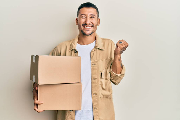 Handsome man with tattoos holding delivery package screaming proud, celebrating victory and success very excited with raised arm  - Photo, Image
