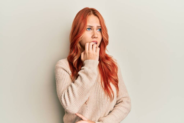 Молодая красивая рыжая женщина в обычном зимнем свитере выглядит напряженной и нервной с руками на рту грызущими ногти. тревожные проблемы.  - Фото, изображение