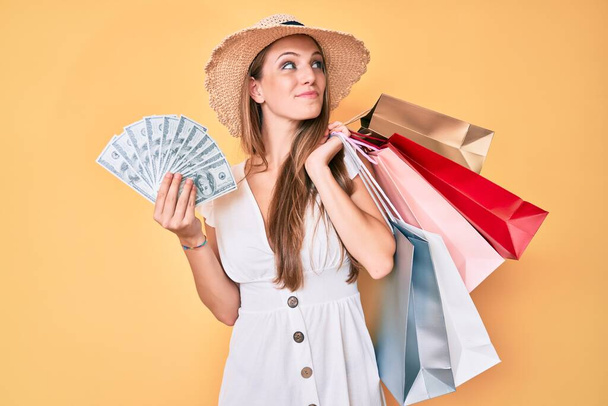 Mladá blondýnka drží nákupní tašky a dolary bankovky s úsměvem dívá do strany a zírá pryč myšlení.  - Fotografie, Obrázek