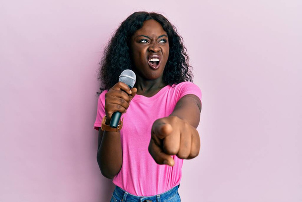 Mooie Afrikaanse jonge vrouw zingen lied met behulp van microfoon wijzen naar camera boos en gek schreeuwen gefrustreerd en woedend, schreeuwen van woede. woede en agressief concept.  - Foto, afbeelding
