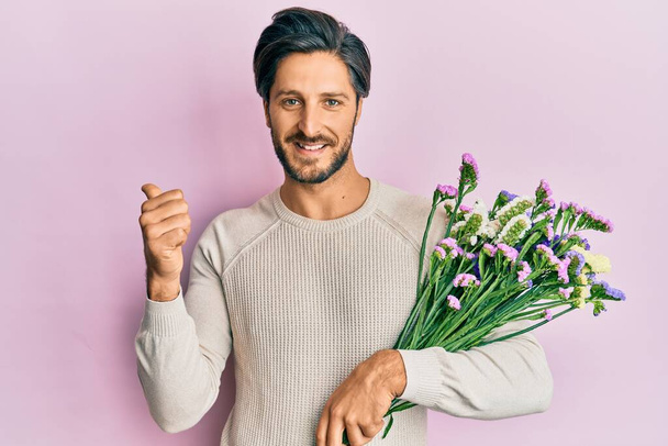 Jeune homme hispanique tenant des fleurs pointant le pouce vers le côté souriant heureux avec la bouche ouverte  - Photo, image