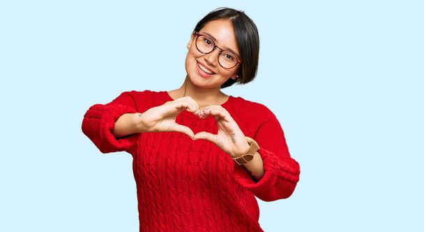 Jonge mooie Latijns-Amerikaanse vrouw met kort haar met casual trui en een bril glimlachend in liefde doet hart symbool vorm met handen. romantisch concept.  - Foto, afbeelding