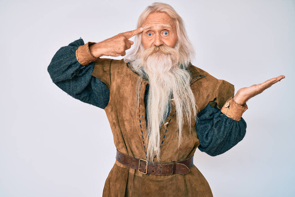 Oude senior man met grijs haar en lange baard dragen viking traditionele kostuum verward en geïrriteerd met open palm tonen kopieerruimte en wijzende vinger naar het voorhoofd. Denk er eens over na..  - Foto, afbeelding