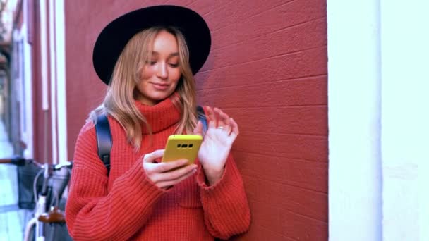 Ξανθιά γυναίκα πληκτρολογώντας και διαβάζοντας μηνύματα κειμένου στο smartphone της μπροστά από έναν κόκκινο τοίχο. - Πλάνα, βίντεο