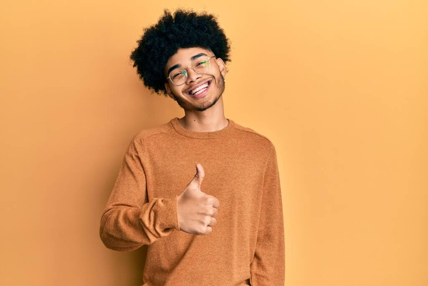 Νεαρός Αφροαμερικάνος με αφρο μαλλιά που φοράει απλό χειμωνιάτικο πουλόβερ και κάνει χαρούμενη χειρονομία με το χέρι. έγκριση έκφρασης κοιτάζοντας την κάμερα που δείχνει την επιτυχία.  - Φωτογραφία, εικόνα