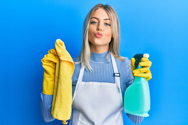 Beautiful blonde woman wearing cleaner apron holding cleaning products looking at the camera blowing a kiss being lovely and sexy. love expression.  - Photo, Image
