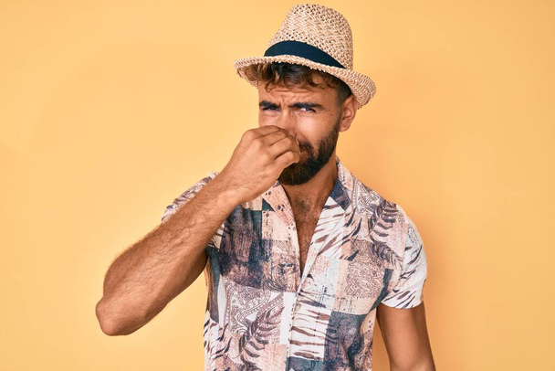 Junger hispanischer Mann mit Sommermütze, der etwas Stinkendes und ekelhaften, unerträglichen Geruch riecht und mit den Fingern auf der Nase den Atem anhält. schlechter Geruch  - Foto, Bild