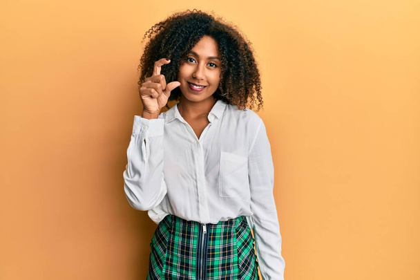 Beautiful african american woman with afro hair wearing scholar skirt smiling and confident gesturing with hand doing small size sign with fingers looking and the camera. measure concept.  - Foto, Imagem