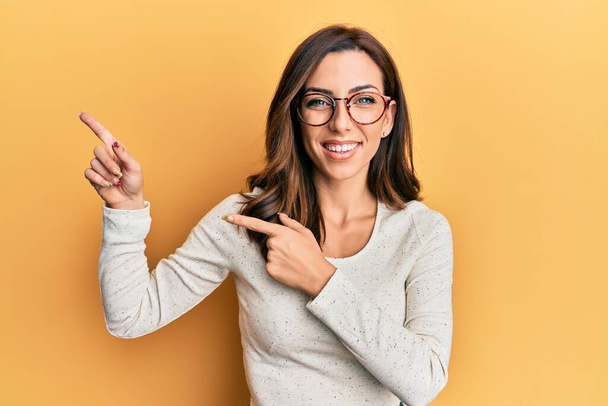Young brunette woman wearing casual clothes and glasses smiling and looking at the camera pointing with two hands and fingers to the side.  - Photo, image
