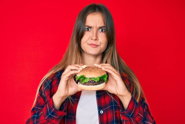 Schöne kaukasische Frau isst einen leckeren klassischen Burger skeptisch und nervös, Stirnrunzeln aufgeregt wegen des Problems. negative person.  - Foto, Bild