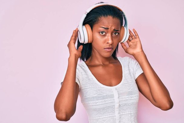 Young african american woman listening to music using headphones skeptic and nervous, frowning upset because of problem. negative person.  - Photo, image