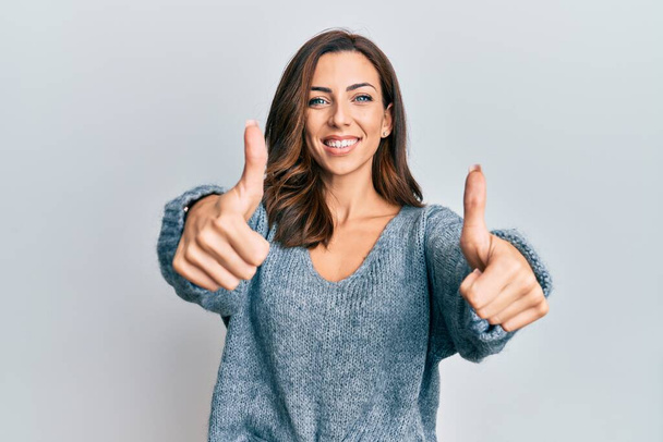 Jeune femme brune portant un pull d'hiver décontracté approuvant faire un geste positif avec la main, les pouces levés souriant et heureux pour le succès. geste gagnant.  - Photo, image