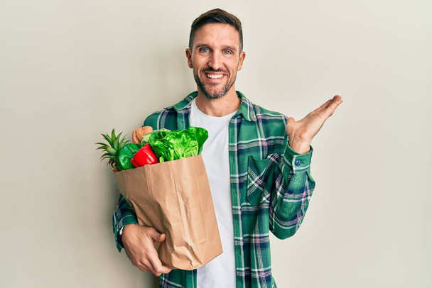 Handsome man with beard holding paper bag with groceries smiling cheerful presenting and pointing with palm of hand looking at the camera.  - Photo, Image