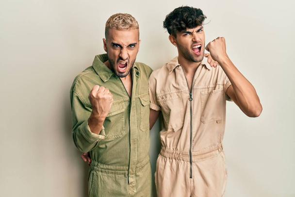 Homosexual gay couple standing together wearing casual jumpsuit angry and mad raising fist frustrated and furious while shouting with anger. rage and aggressive concept.  - Foto, immagini