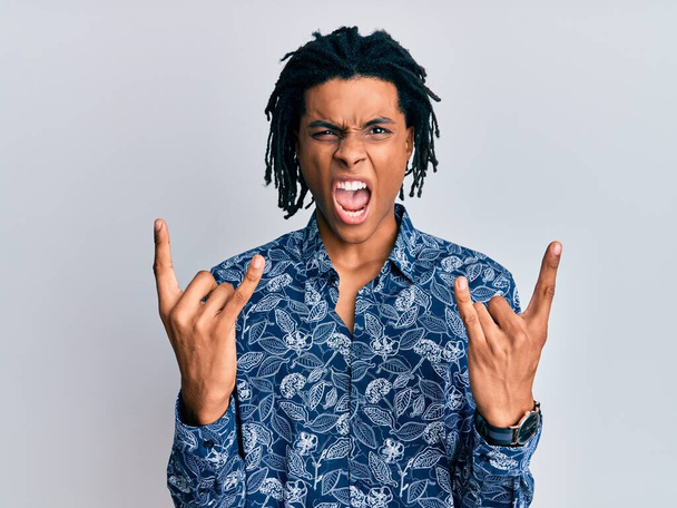 Joven hombre afroamericano con camisa de estilo 80 gritando con expresión loca haciendo símbolo de rock con las manos arriba. Estrella de la música. concepto pesado.  - Foto, imagen
