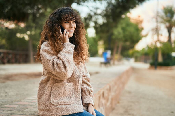 Jovem hispânica sorrindo feliz falando no smartphone no parque. - Foto, Imagem