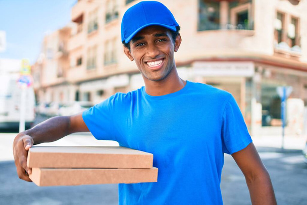 Kurye üniforması giyen Afrikalı teslimatçı dışarıda pizza kutularını tutuyor. - Fotoğraf, Görsel