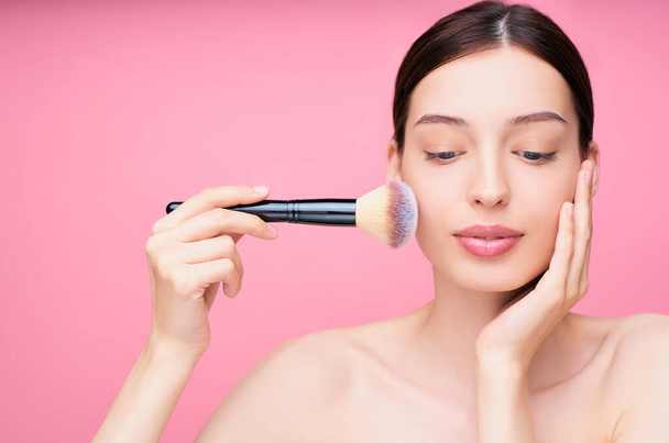Beautiful brown-haired millennial girl doing makeup, applying powder with a cosmetic brush, looking down, posing on a pink studio background. Makeup advertising banner with free space. Copy space. - Foto, Imagen