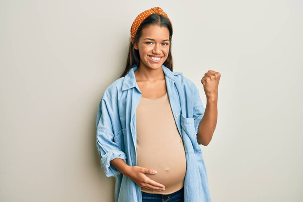 Beautiful hispanic woman expecting a baby, touching pregnant belly very happy and excited doing winner gesture with arms raised, smiling and screaming for success. celebration concept.  - Fotó, kép