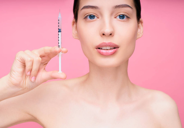 Close-up, Skin lifting injection. Young attractive charming caucasian woman holding a syringe with a medicine in her hand, standing on a pink background. Studio . - Photo, image
