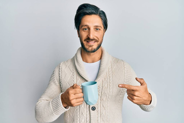 Nuori latino mies juo kupin kahvia hymyillen onnellinen osoittaa kädellä ja sormella  - Valokuva, kuva