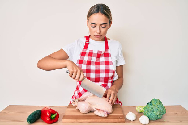 Young beautiful blonde woman wearing cook apron cutting chicken relaxed with serious expression on face. simple and natural looking at the camera.  - Foto, Imagen
