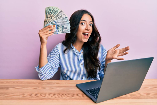Красивая брюнетка молодая женщина, работающая в офисе, держа в руках доллары, празднуя достижение с счастливой улыбкой и выражением победителя с поднятой рукой  - Фото, изображение