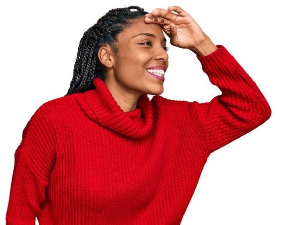 Afroamerikanerin in lässigem Winterpullover sehr glücklich und lächelnd, die weit weg mit der Hand über dem Kopf schaut. Suchkonzept.  - Foto, Bild