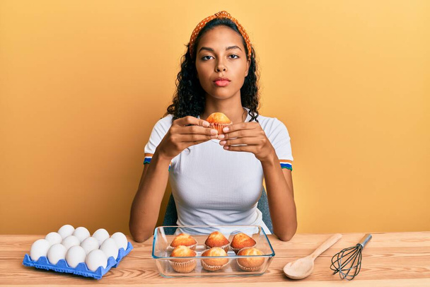 Молодая африканская американская девушка делает кексы сидя на столе мышление отношение и трезвое выражение лица, глядя самоуверенно  - Фото, изображение