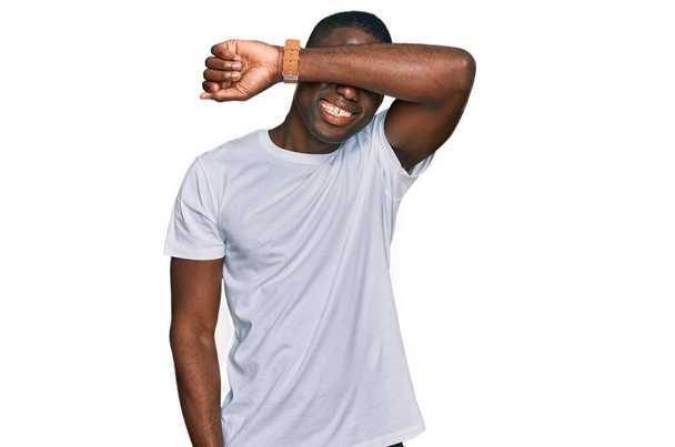 Mladý africký Američan v neformální bílé tričko zakrývající oči s úsměvem na rameni vesele a legračně. slepý koncept.  - Fotografie, Obrázek
