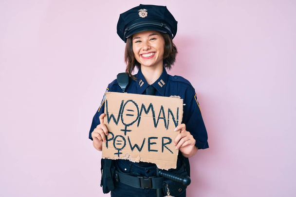 Молодая красивая девушка в полицейской форме, держащая женское знамя власти выглядит позитивно и счастливо стоя и улыбаясь с уверенной улыбкой, показывая зубы  - Фото, изображение