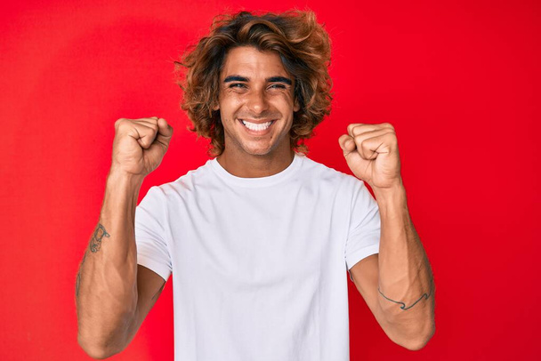 Молодий іспаномовний чоловік в повсякденній білій сорочці кричить гордо, відзначаючи перемогу і успіх дуже схвильований піднятими руками
  - Фото, зображення