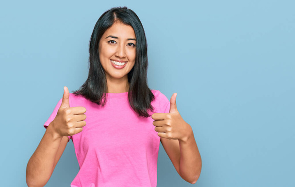 Bella asiatica giovane donna indossa casual t shirt rosa approvazione facendo gesto positivo con la mano, pollice in su sorridente e felice per il successo. gesto vincitore.  - Foto, immagini