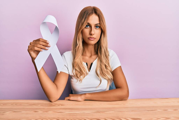 Beautiful blonde young woman holding white ribbon thinking attitude and sober expression looking self confident  - Foto, Imagen